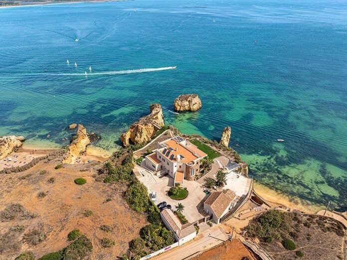 Cenário natural à beira-mar com vista para o horizonte, apresentando águas azuis e uma propriedade elegante rodeada por vegetação.