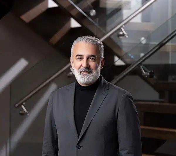 Homem de cabelo grisalho, barba bem cuidada, vestido em um ambiente elegante e contemporâneo, sentado em um sofá moderno com detalhes em vidro e metal.