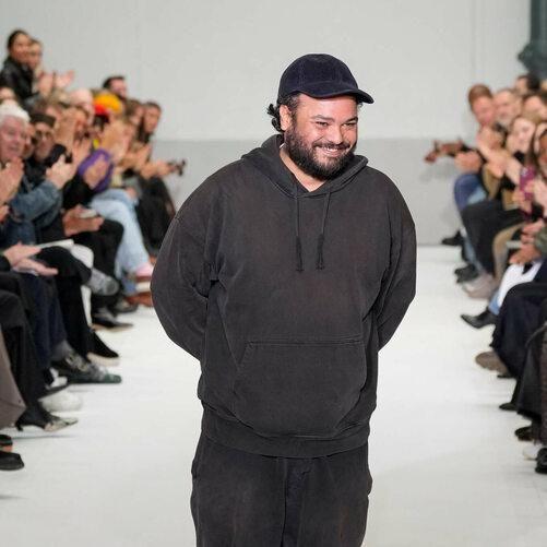 Homem no desfile, usando moletom escuro, sorridente, com plateia ao fundo aplaudindo.
