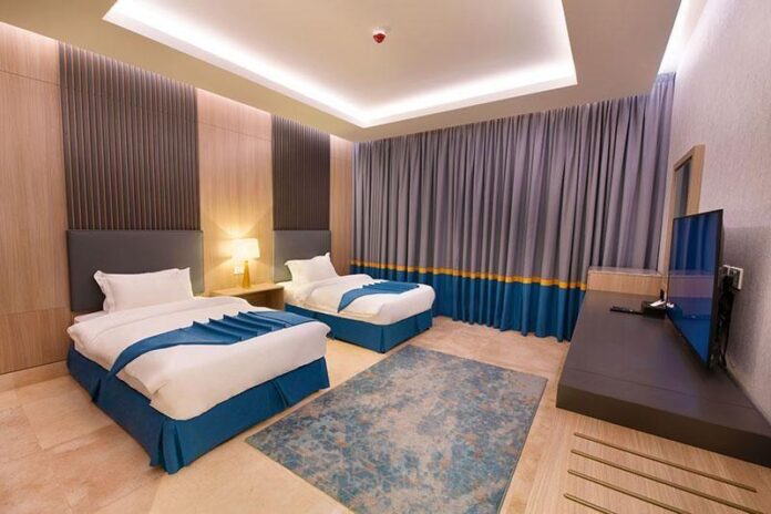 Quarto com paredes de madeira e cortinas em tons de cinza e azul, camas com lençóis brancos, luz suave e uma televisão na parede.