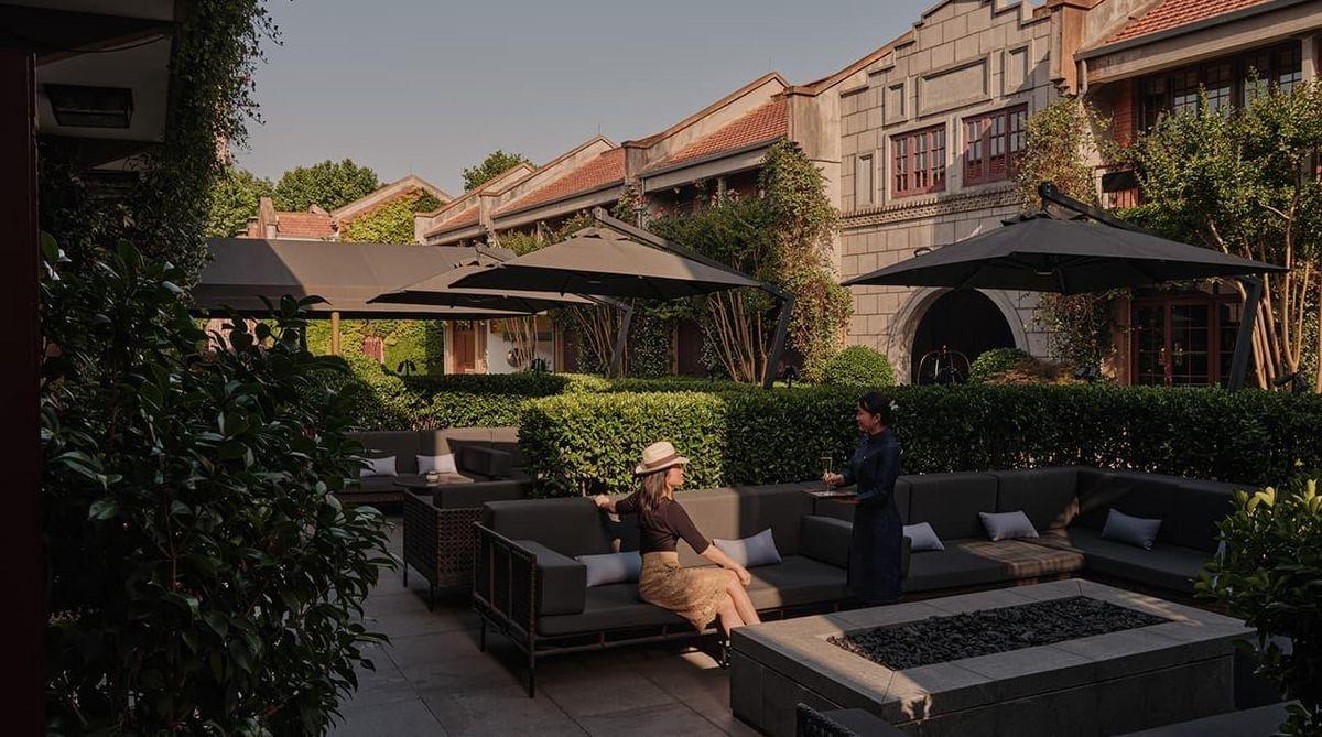 Área externa do hotel Capella Shanghai, com sofás e guarda-sóis, cercada por edifícios de arquitetura histórica.