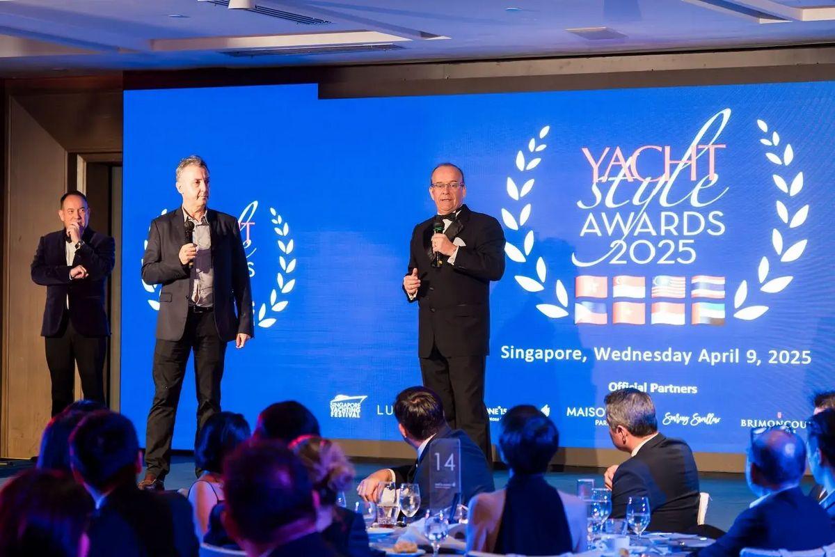 Homens de terno falando ao microfone em um palco durante o Yacht Style Awards 2025, com uma tela azul ao fundo com logotipo do evento.
