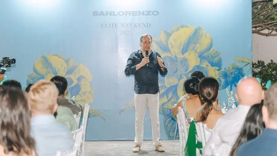 Massimo Perotti em pé, segurando um microfone, frente a uma plateia em um evento, com um fundo floral em azul e amarelo.