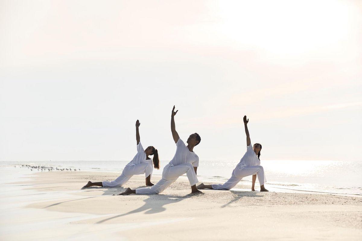 Três praticantes de yoga na praia com o mar ao fundo e céu claro