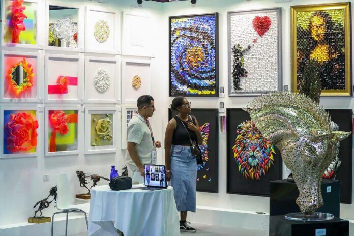 Um ambiente de exposição com paredes brancas, quadros emoldurados e duas pessoas observando as obras.