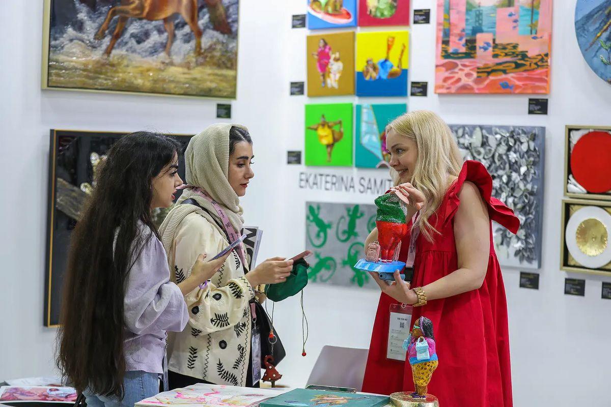 Um fundo branco com várias obras de arte expostas nas paredes, mulheres com roupas variadas e estatuetas coloridas.