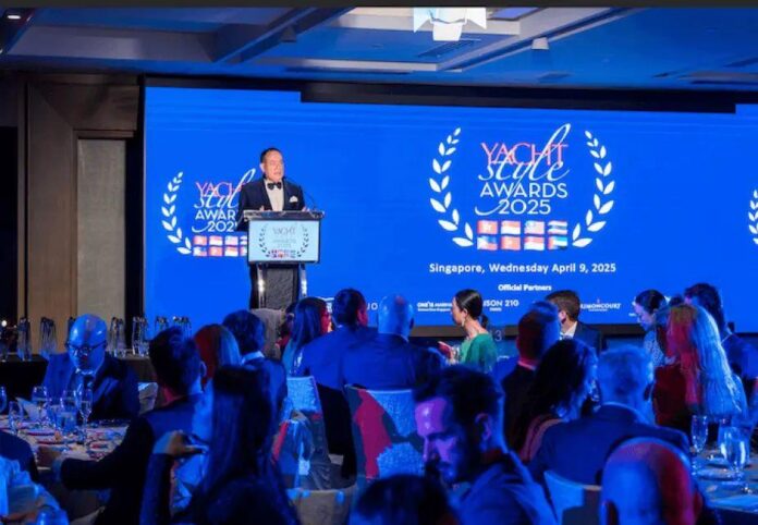 Um homem em terno segurando um microfone durante a apresentação no Yacht Style Awards 2025, com um fundo azul claro e logotipo do evento.